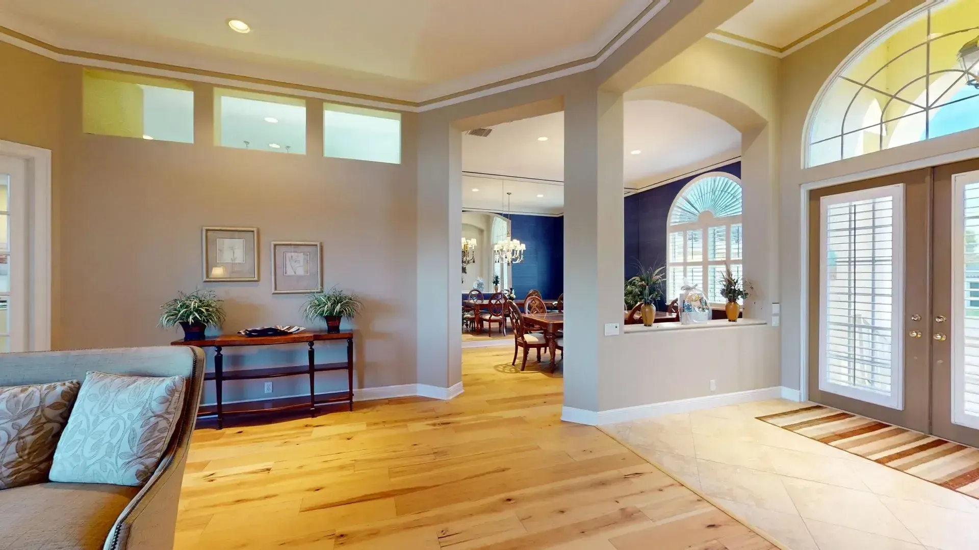 Spacious living room with light hardwood floors, high ceilings, a tan sofa and armchair, and large glass sliding doors that open to a covered patio with a waterfront view.