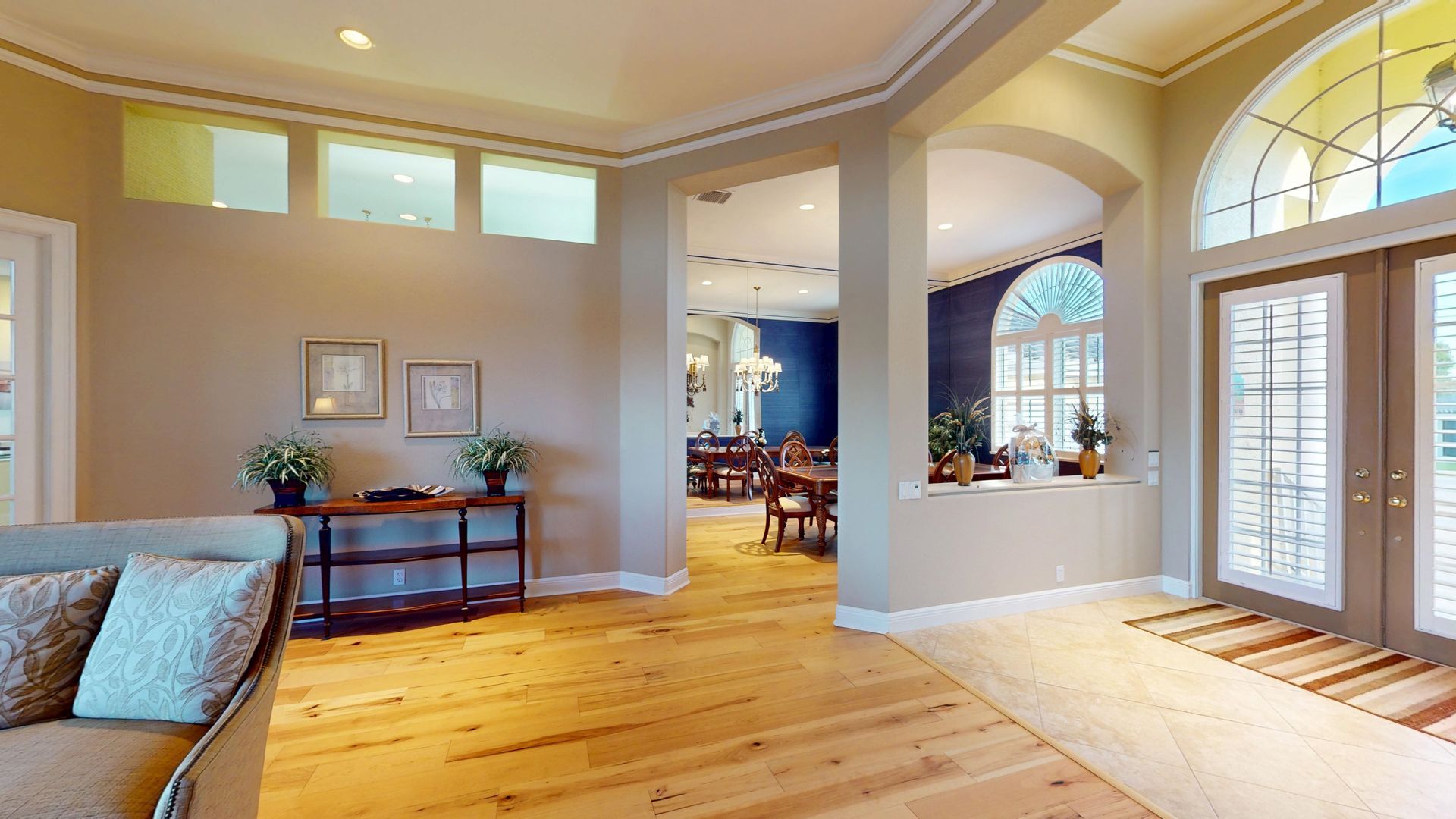 Spacious living room with light hardwood floors, high ceilings, a tan sofa and armchair, and large glass sliding doors that open to a covered patio with a waterfront view.