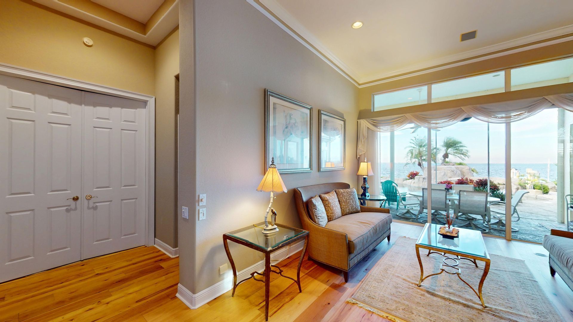 Bright, elegantly decorated living room featuring a neutral color palette, twin framed floral artwork over a classic sofa, warm accent lighting, and elegant window valences over glass doors.