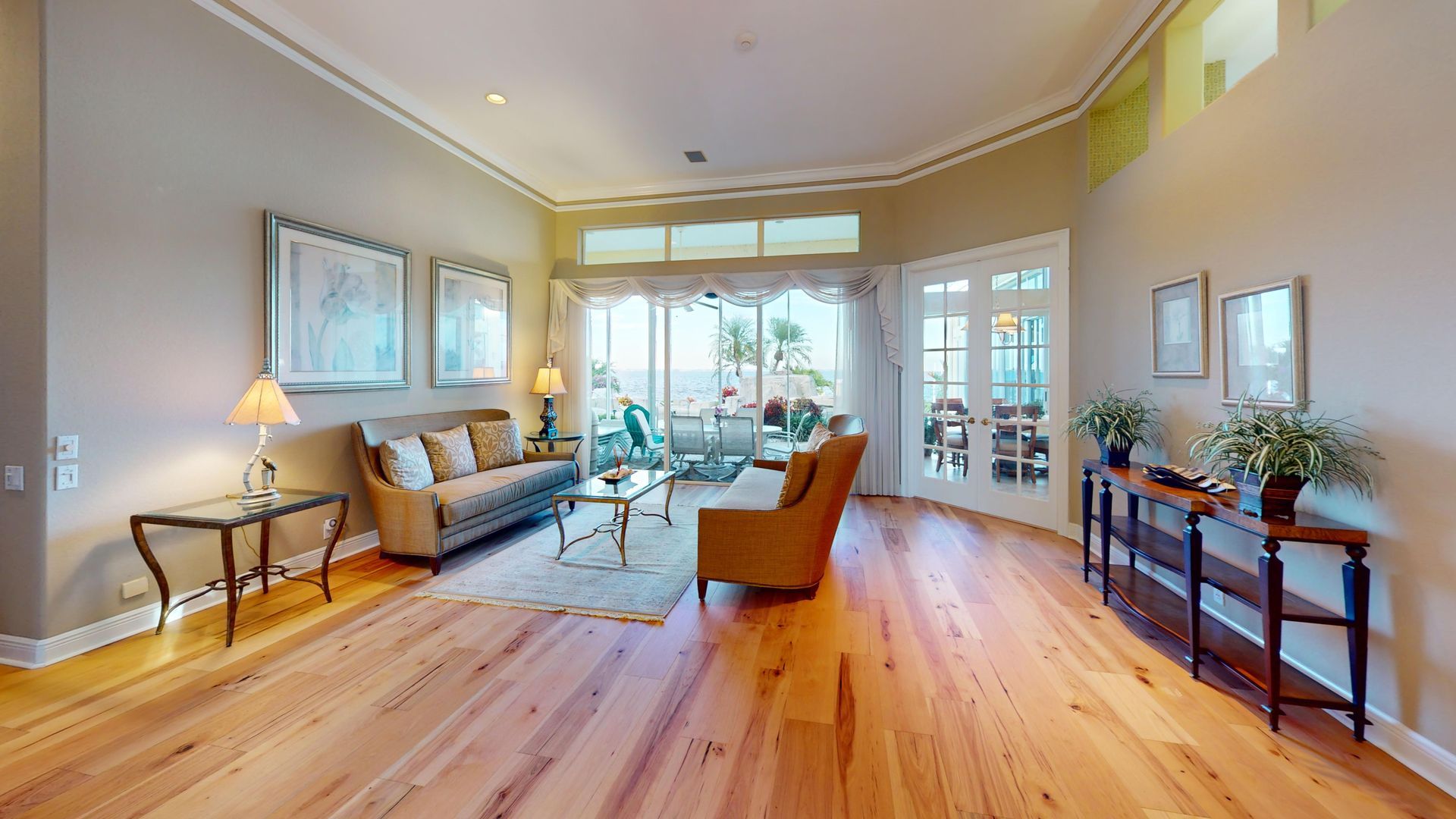 A brightly lit, upscale living room with hardwood floors, comfortable seating, and large glass doors leading to an outdoor patio with water views.