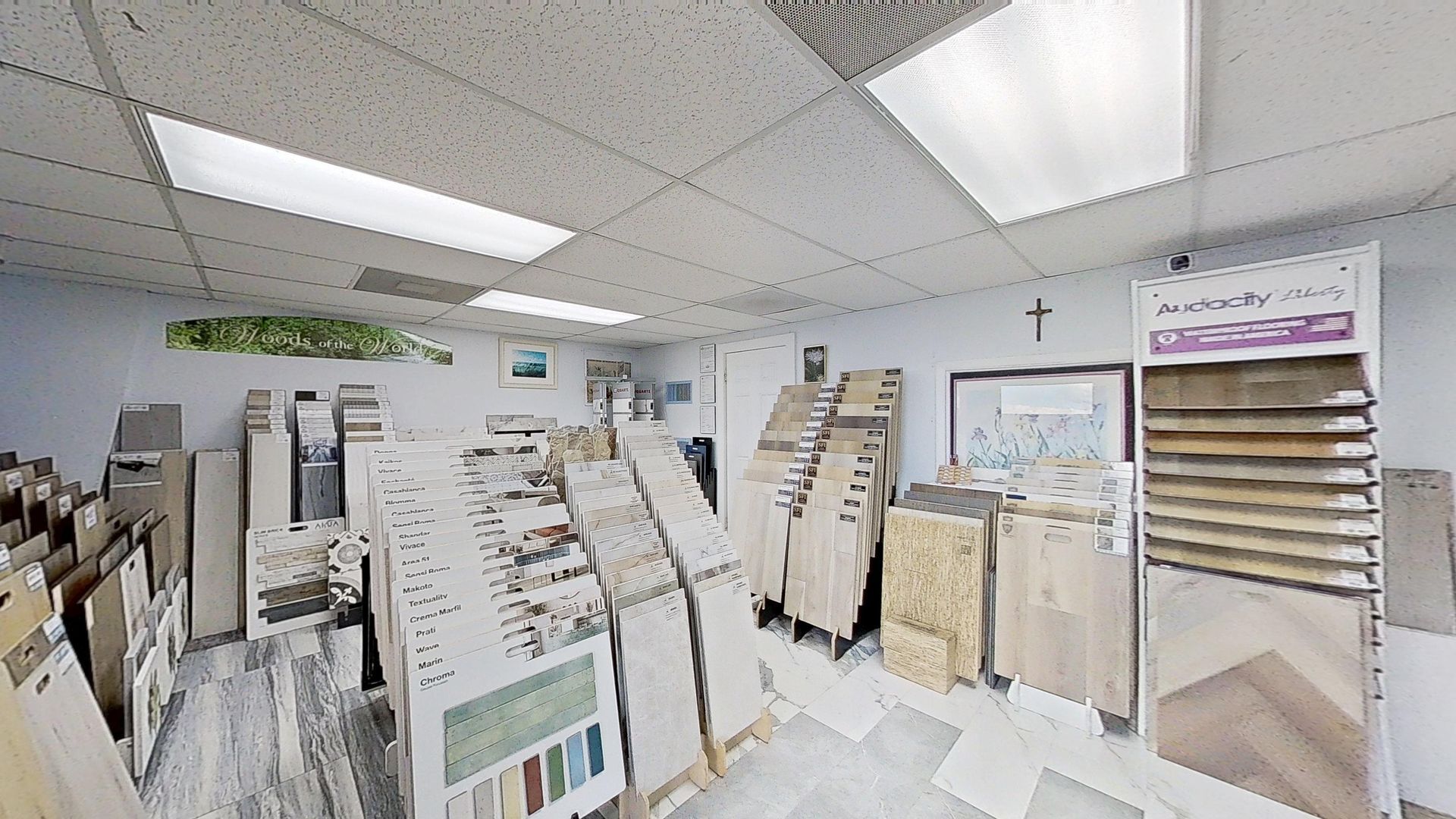 An indoor showroom wall displaying rows of hanging mosaic and tile samples in various geometric shapes, colors, and textures, next to an Audacity brand flooring display.