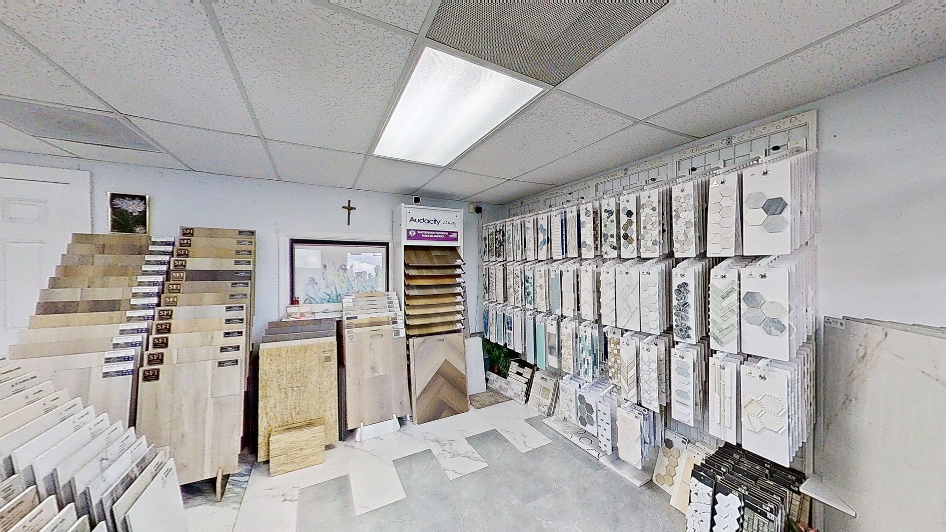 An indoor showroom wall displaying rows of hanging mosaic and tile samples in various geometric shapes, colors, and textures, next to an Audacity brand flooring display.