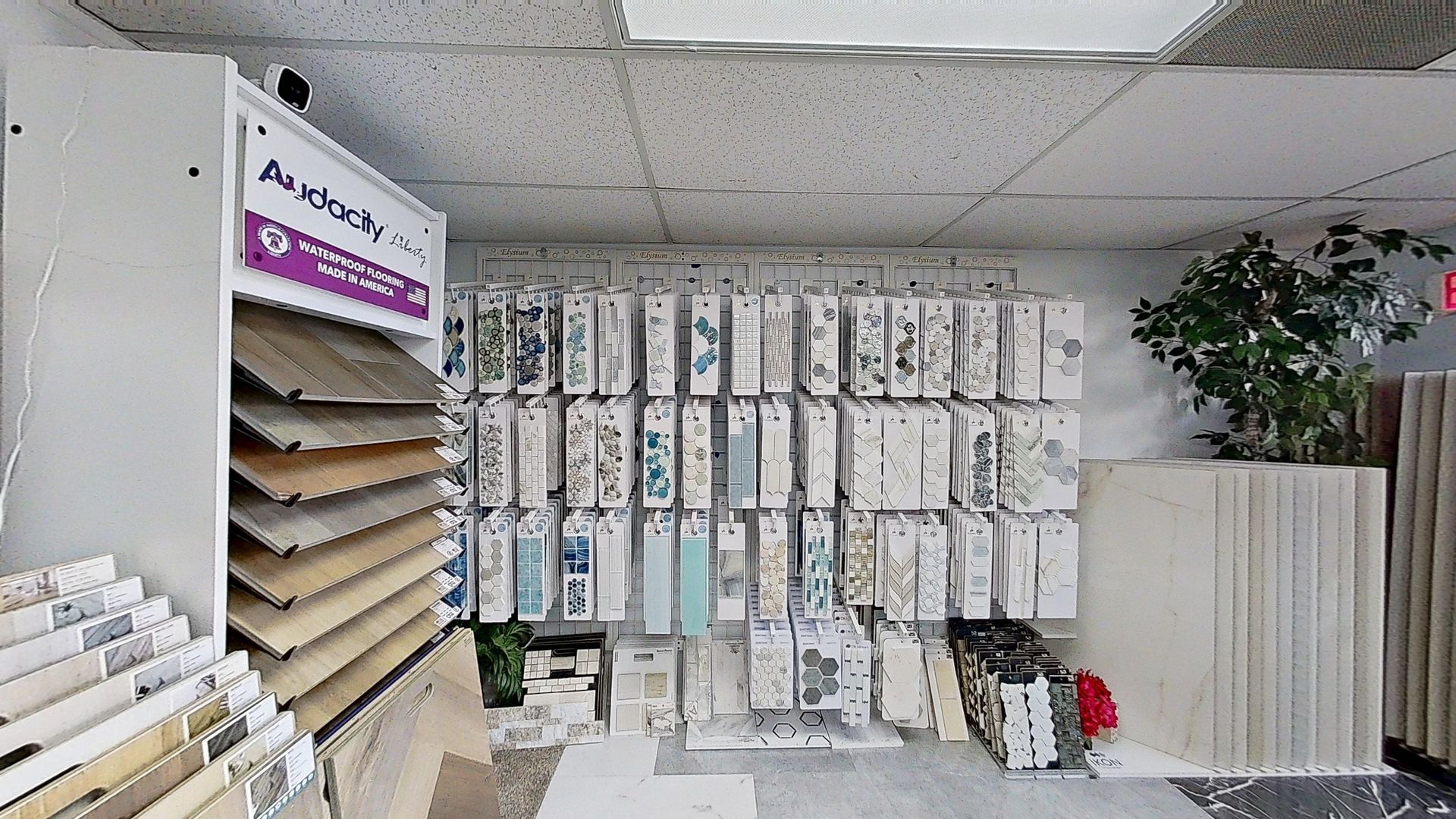 An indoor showroom wall displaying rows of hanging mosaic and tile samples in various geometric shapes, colors, and textures, next to an Audacity brand flooring display.