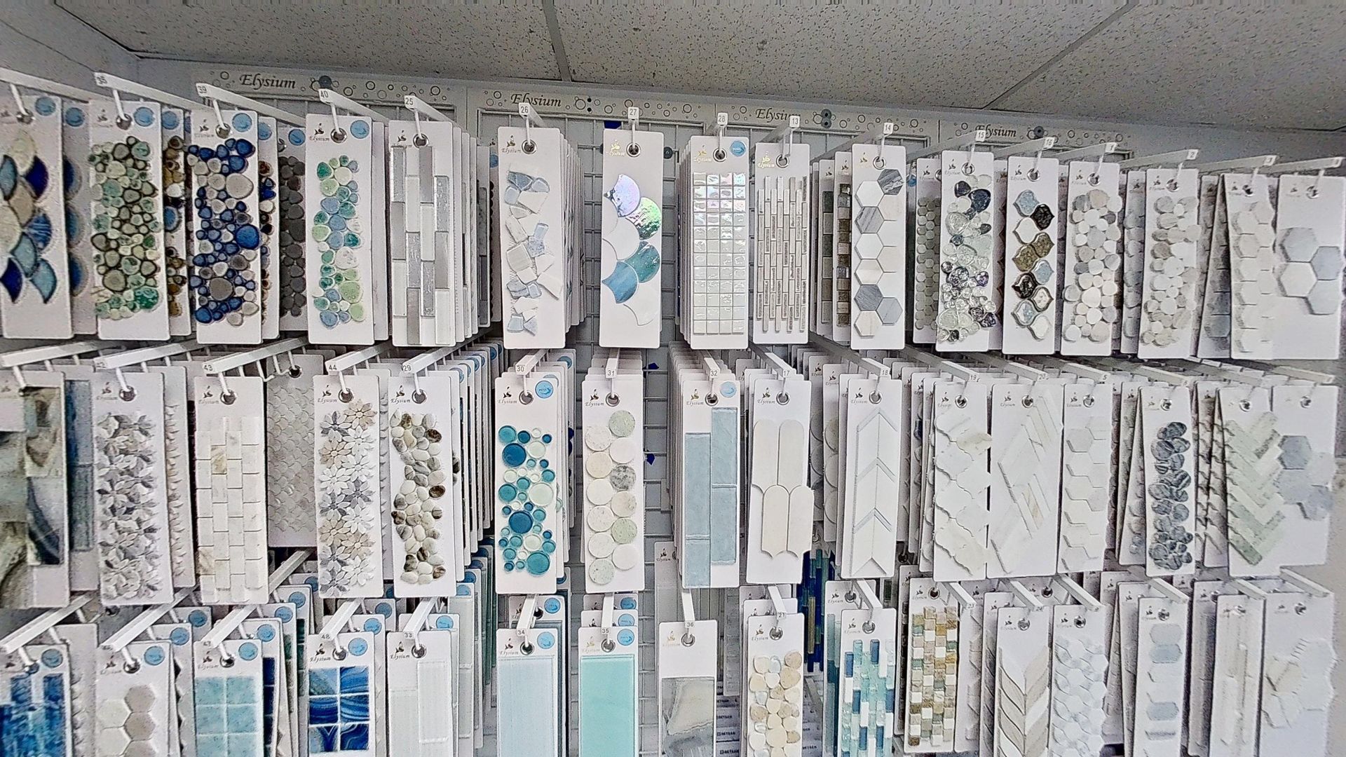 An indoor showroom wall displaying rows of hanging mosaic and tile samples in various geometric shapes, colors, and textures, next to an Audacity brand flooring display.