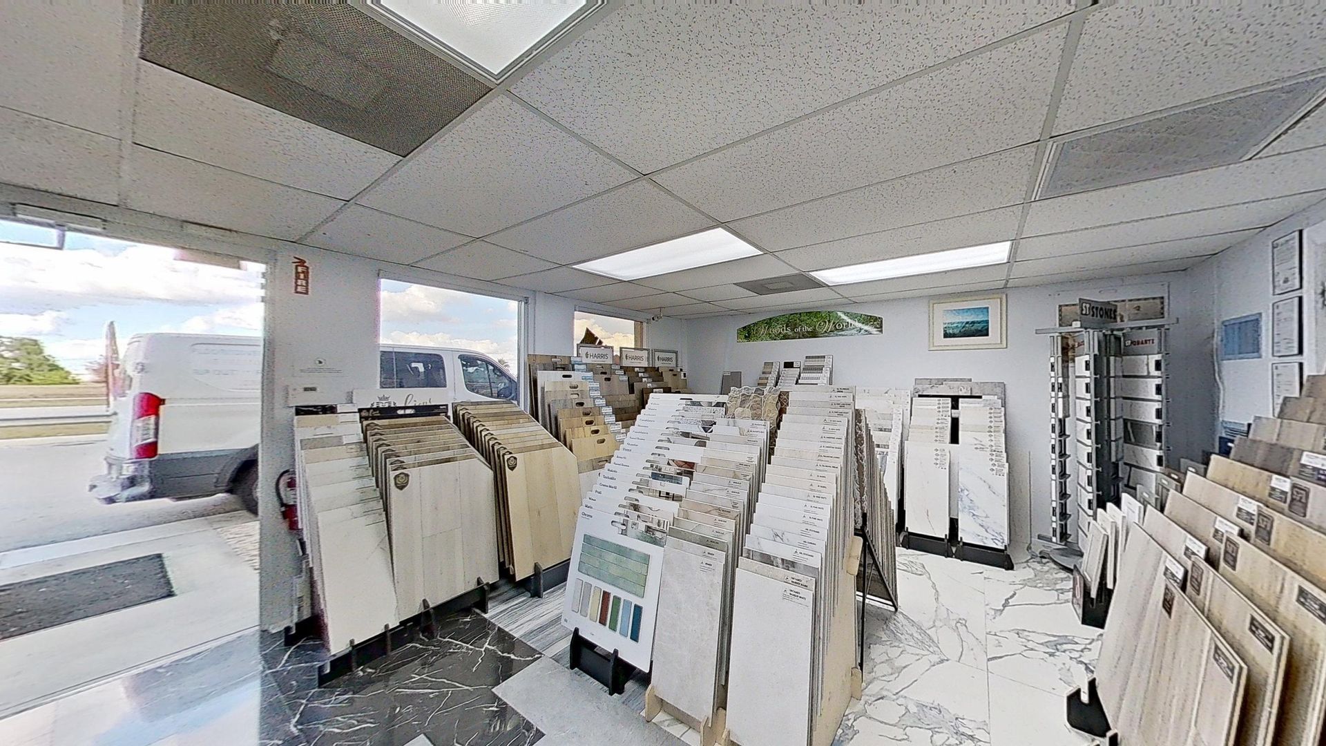 An indoor showroom wall displaying rows of hanging mosaic and tile samples in various geometric shapes, colors, and textures, next to an Audacity brand flooring display.