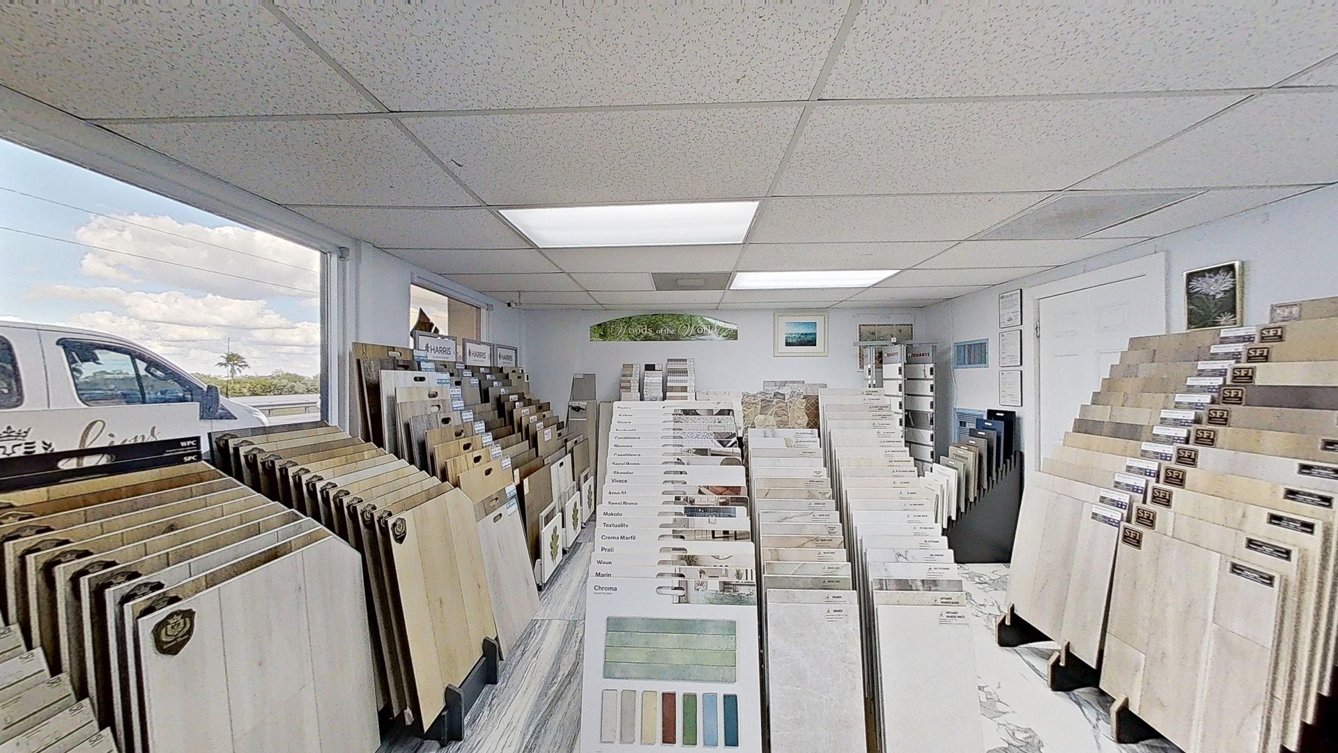 An indoor showroom wall displaying rows of hanging mosaic and tile samples in various geometric shapes, colors, and textures, next to an Audacity brand flooring display.