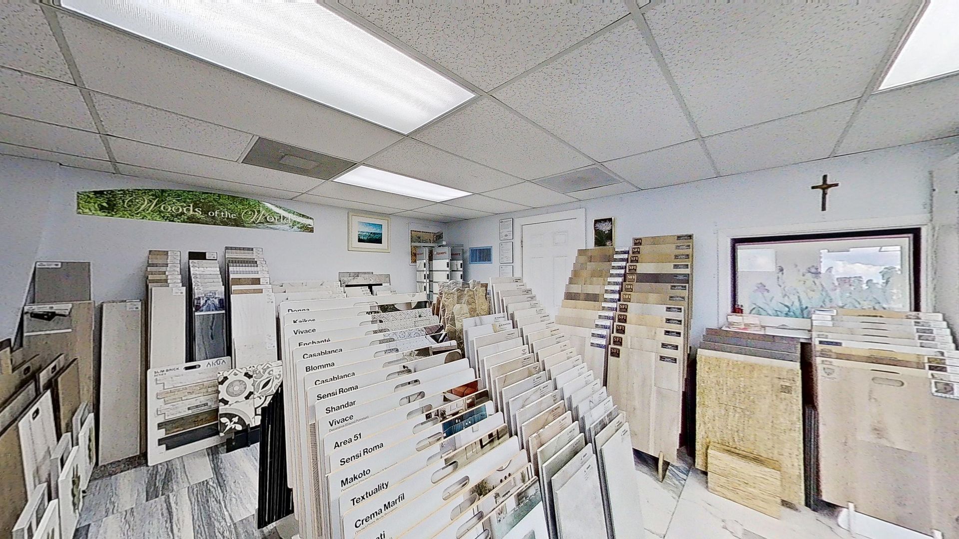 An indoor showroom wall displaying rows of hanging mosaic and tile samples in various geometric shapes, colors, and textures, next to an Audacity brand flooring display.