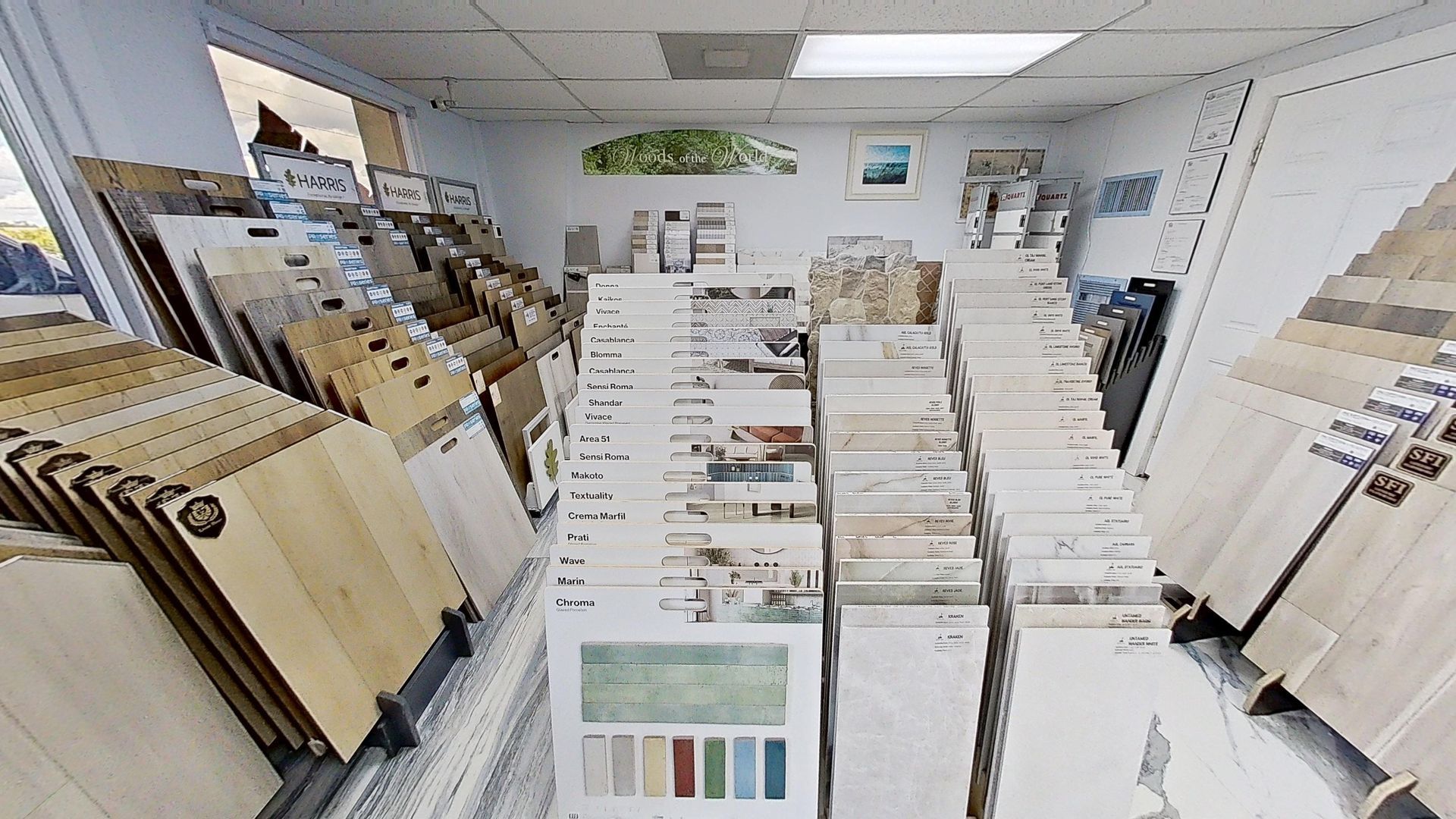 An indoor showroom wall displaying rows of hanging mosaic and tile samples in various geometric shapes, colors, and textures, next to an Audacity brand flooring display.