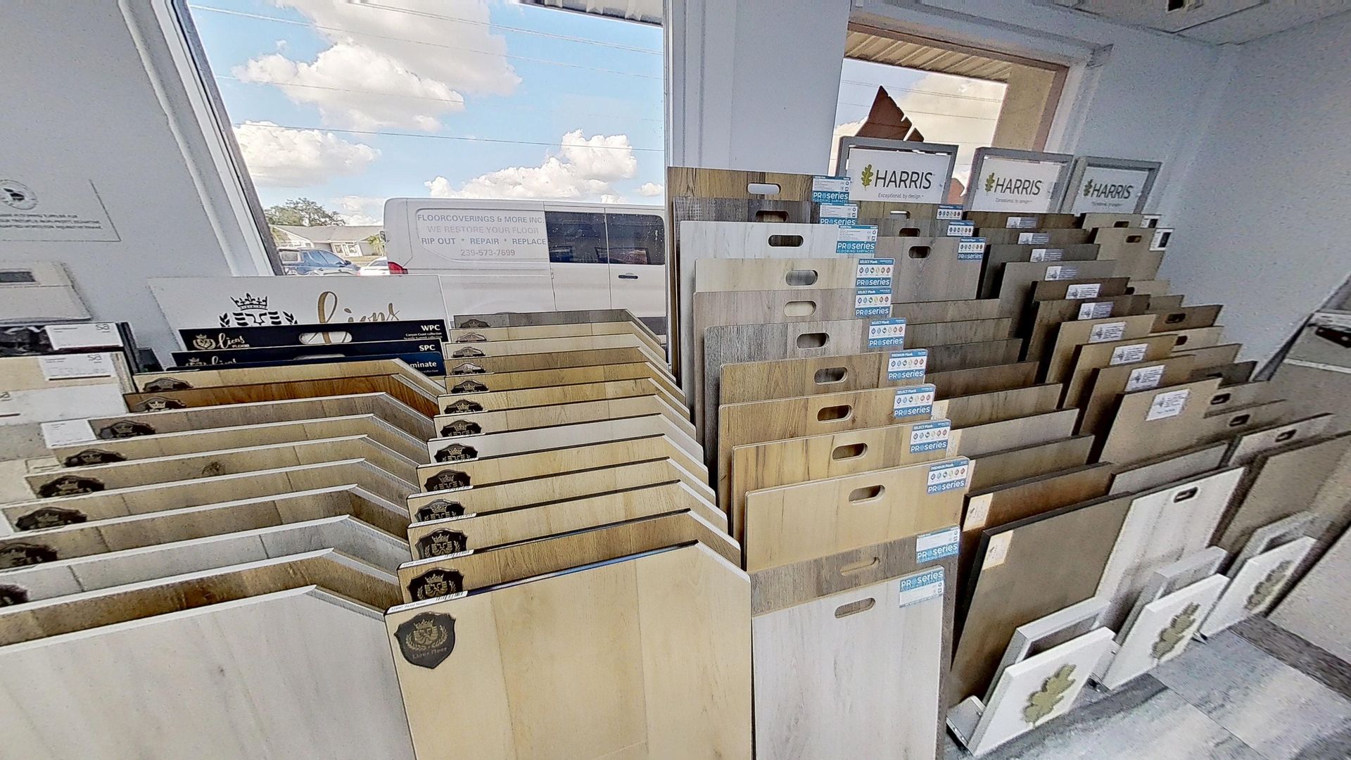 An indoor showroom wall displaying rows of hanging mosaic and tile samples in various geometric shapes, colors, and textures, next to an Audacity brand flooring display.