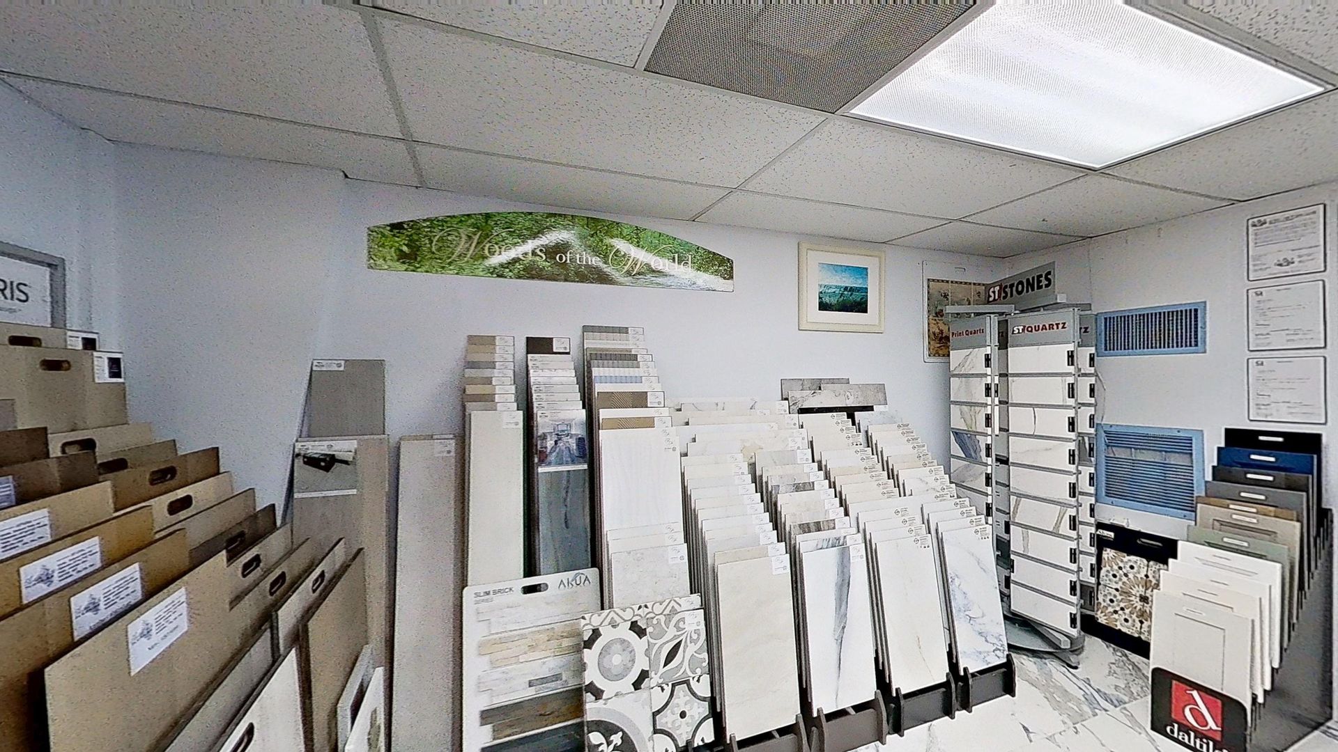 An indoor showroom wall displaying rows of hanging mosaic and tile samples in various geometric shapes, colors, and textures, next to an Audacity brand flooring display.