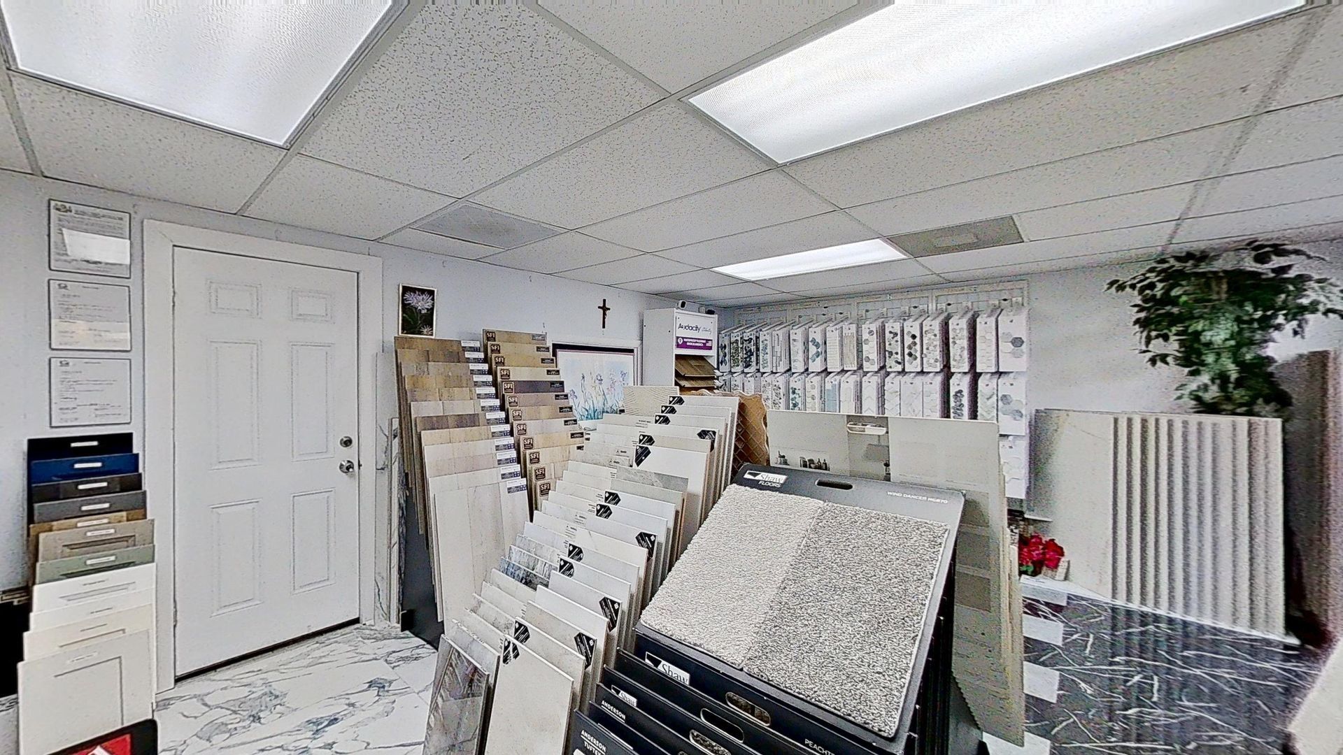 An indoor showroom wall displaying rows of hanging mosaic and tile samples in various geometric shapes, colors, and textures, next to an Audacity brand flooring display.