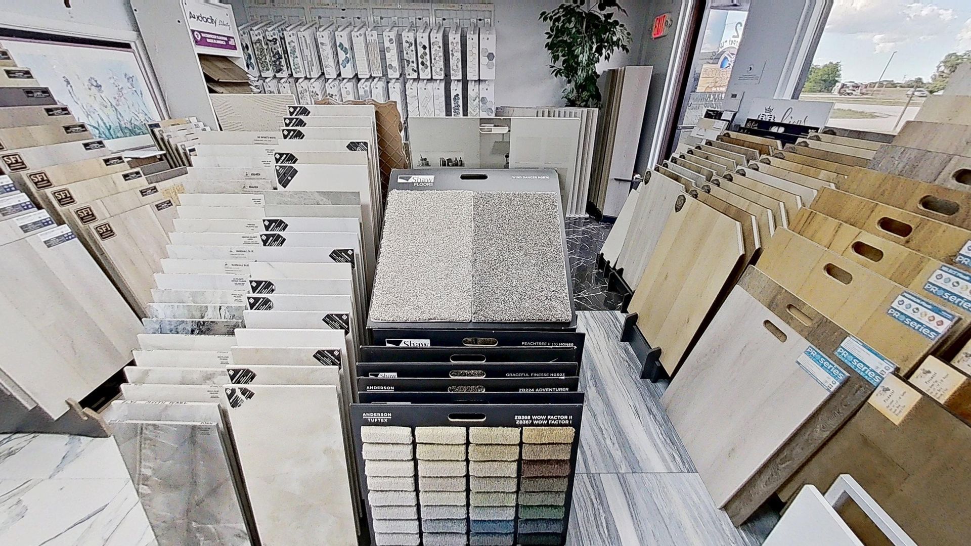 An indoor showroom wall displaying rows of hanging mosaic and tile samples in various geometric shapes, colors, and textures, next to an Audacity brand flooring display.