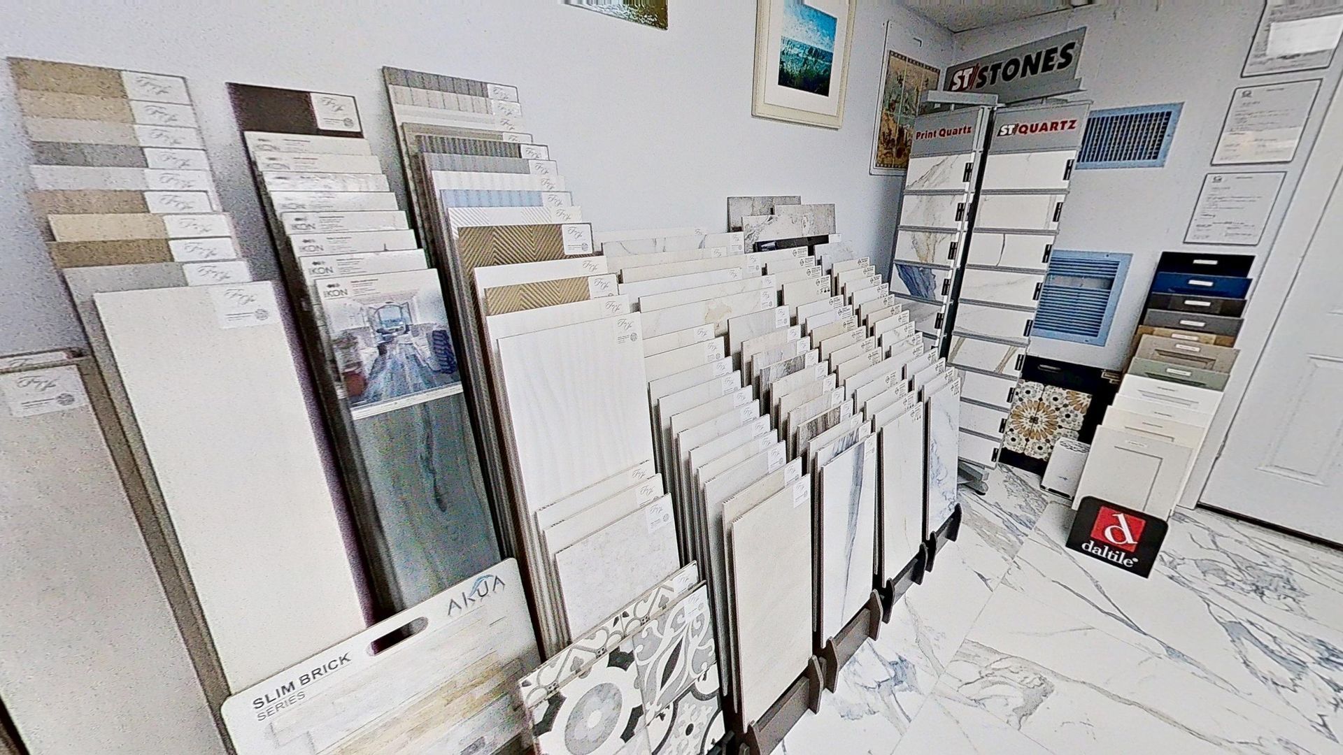An indoor showroom wall displaying rows of hanging mosaic and tile samples in various geometric shapes, colors, and textures, next to an Audacity brand flooring display.