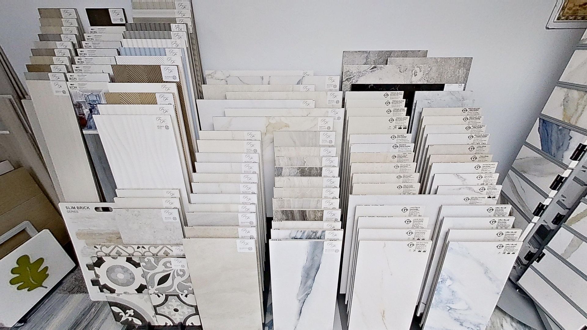An indoor showroom wall displaying rows of hanging mosaic and tile samples in various geometric shapes, colors, and textures, next to an Audacity brand flooring display.