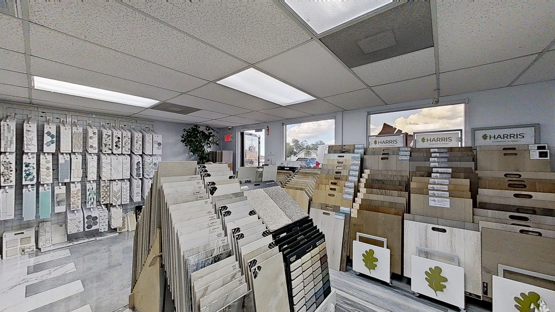 An indoor showroom wall displaying rows of hanging mosaic and tile samples in various geometric shapes, colors, and textures, next to an Audacity brand flooring display.
