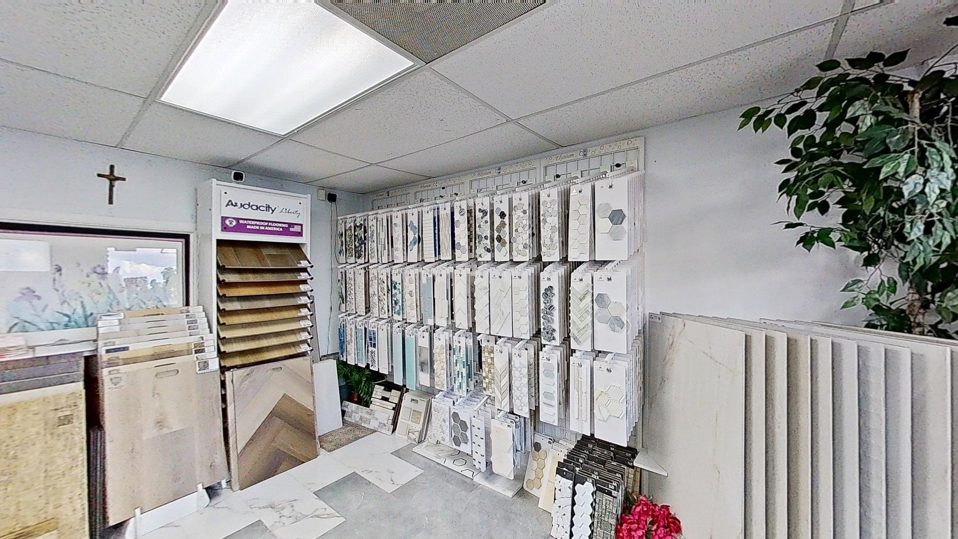 An indoor showroom wall displaying rows of hanging mosaic and tile samples in various geometric shapes, colors, and textures, next to an Audacity brand flooring display.