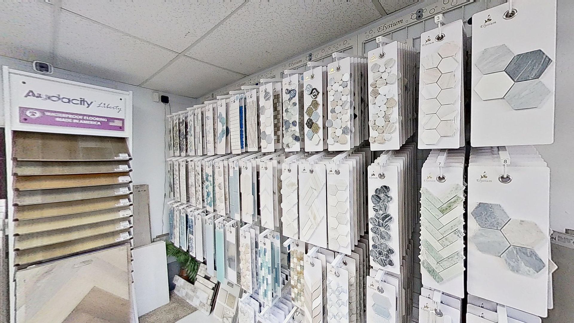 An indoor showroom wall displaying rows of hanging mosaic and tile samples in various geometric shapes, colors, and textures, next to an Audacity brand flooring display.