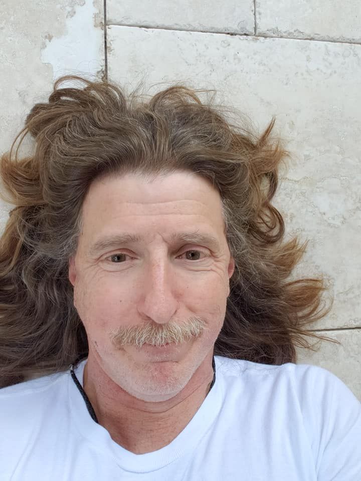 Man with long, wavy brown hair and mustache lying on a light-colored tile surface, wearing a white t-shirt.