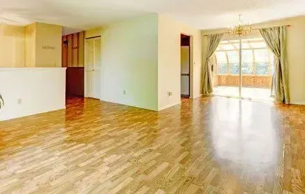 Empty room with wood floors and sliding glass doors to a sunroom, with tan walls and curtains.
