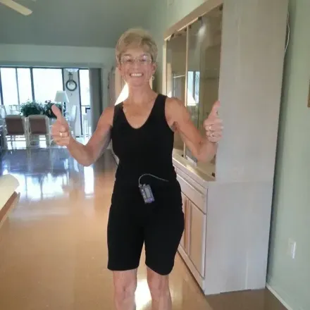 Woman in black workout clothes giving thumbs up; in a home with a cabinet.