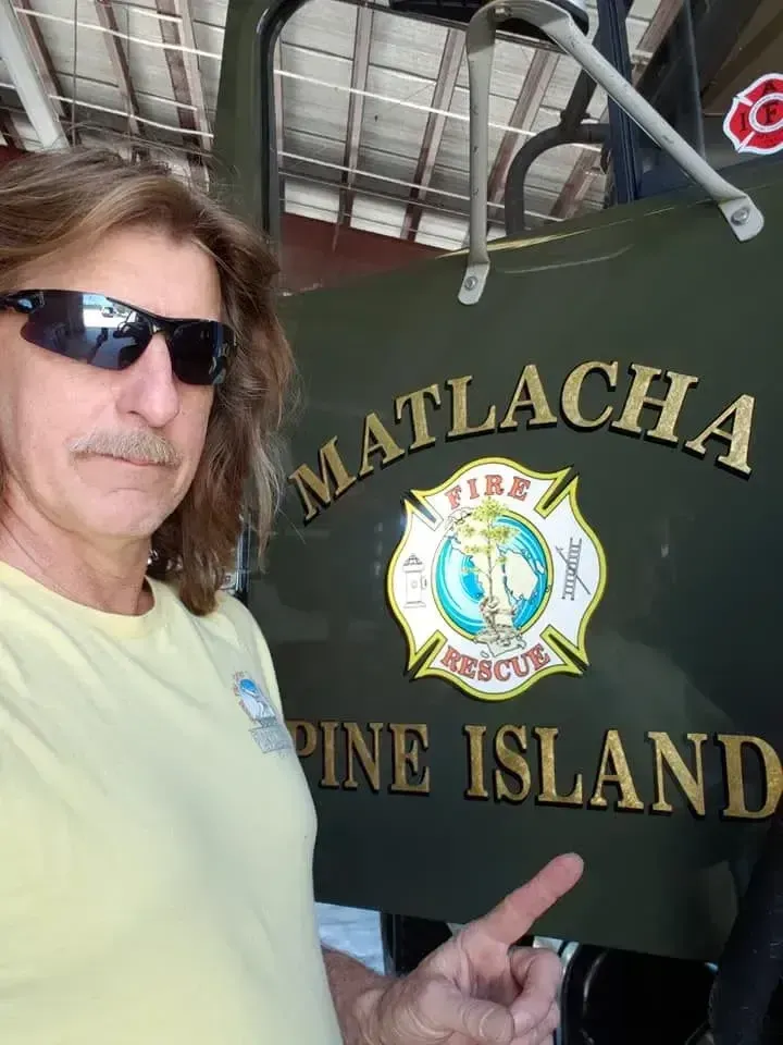A man with long hair and sunglasses pointing to a logo on a green fire truck that reads 'Matlacha Pine Island Fire Rescue'.