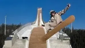 A snowboarder in mid-air performing a trick off a massive, curved ski jump ramp under a clear blue sky.