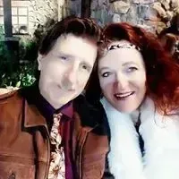 A close-up selfie of a smiling couple indoors. The man wears a brown leather jacket and patterned tie, and the woman has long red hair and a white furry coat.