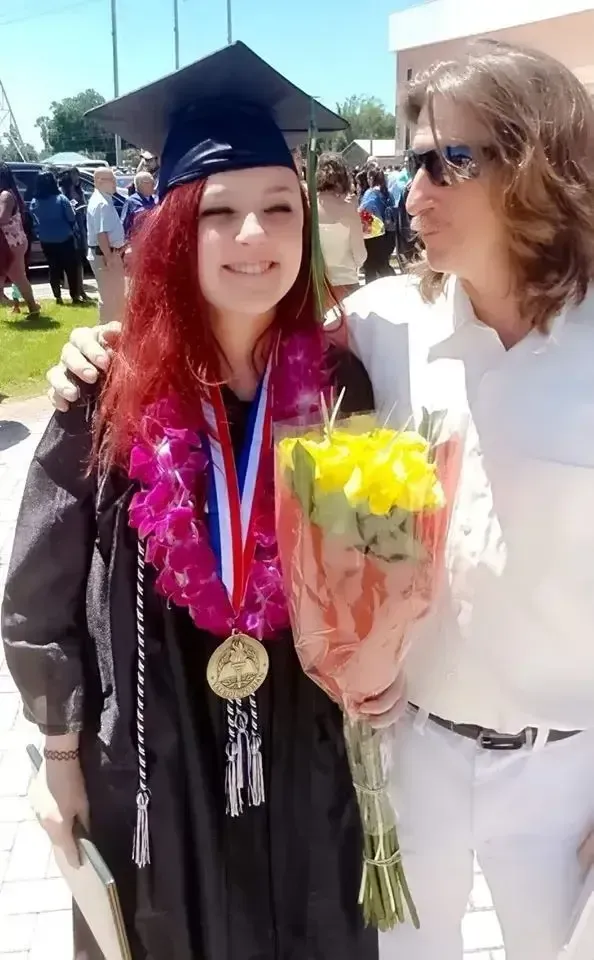 A smiling female graduate with red hair wearing a black cap and gown, a purple flower lei, and a valedictorian medal, standing next to a man with long hair and sunglasses who is holding a bouquet of yellow roses.