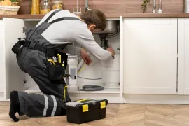 Plumber kneeling under kitchen sink, repairing pipes beside open cabinet with tools nearby.