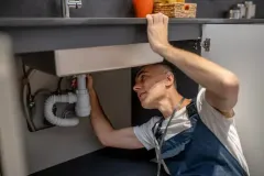 Person repairing plumbing under a sink, reaching up inside the cabinet.