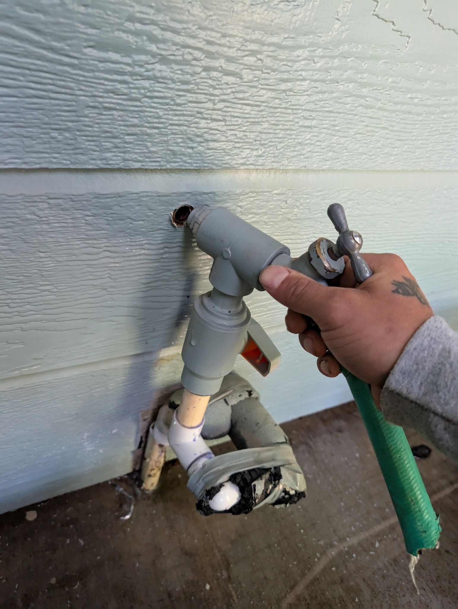 A hand holds a loose, leaking outdoor faucet assembly connected by gray PVC pipes extending from a hole in a house wall.