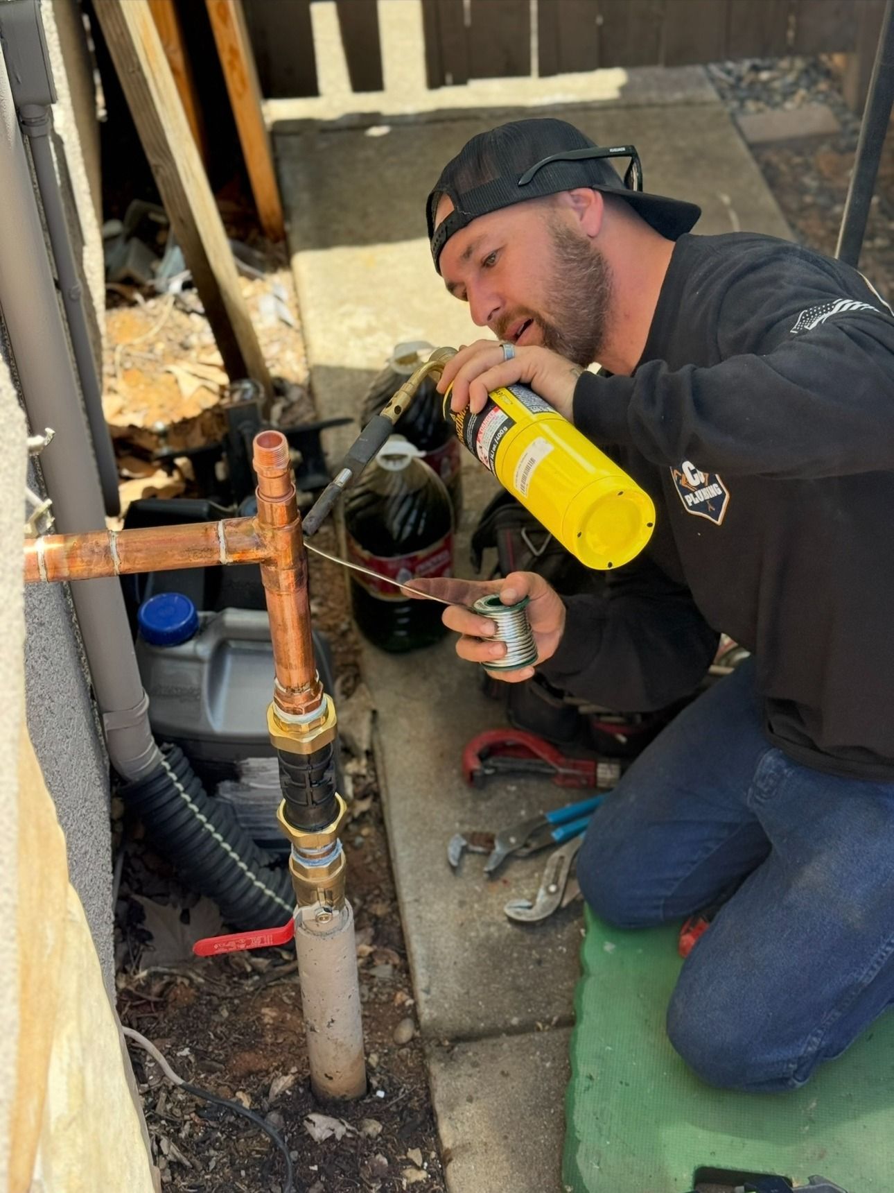 A plumber kneels outdoors, using a yellow torch to solder a copper pipe fitting on a home exterior.