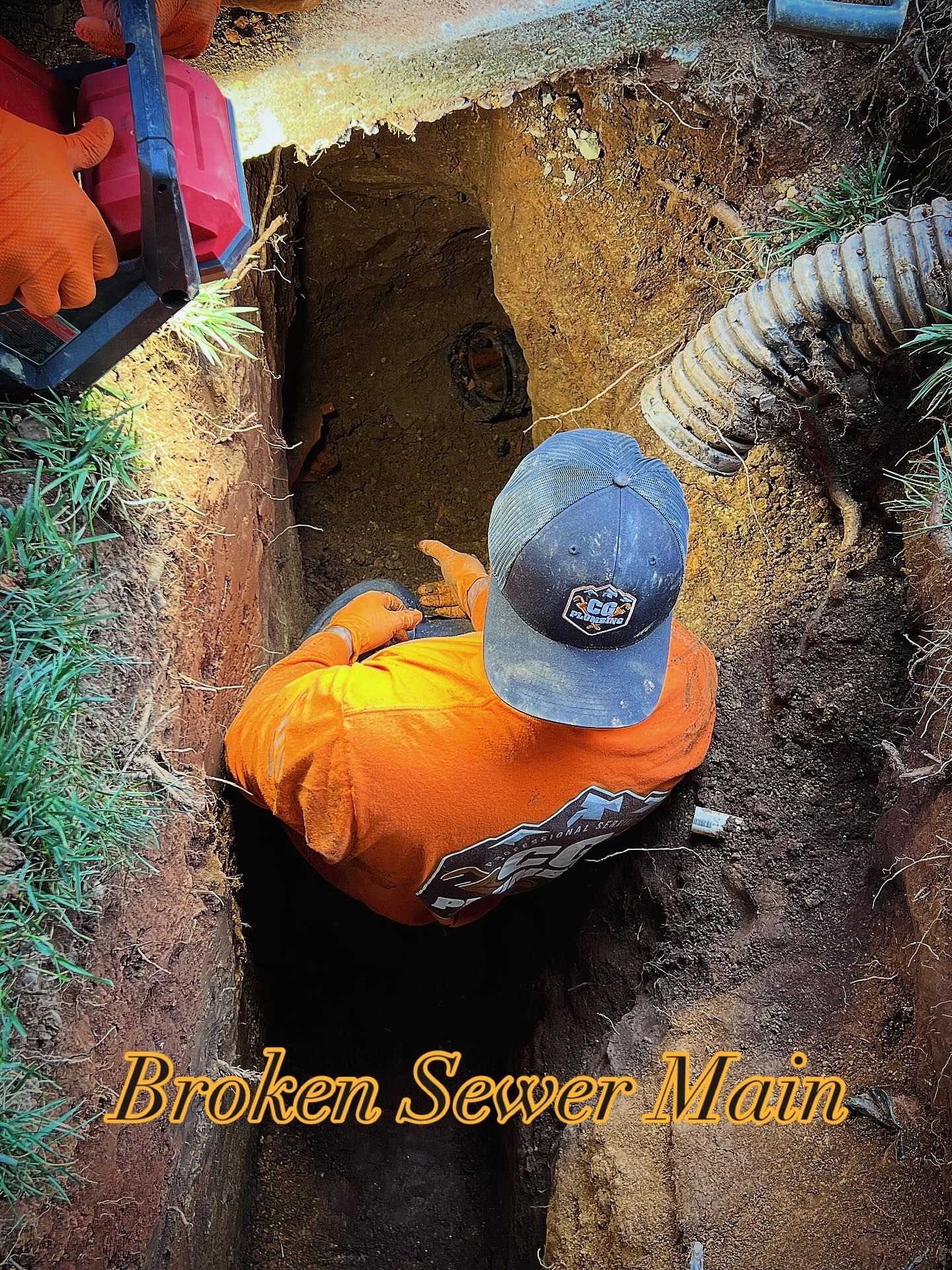 A worker in an orange shirt and cap stands in an excavated trench to repair a broken sewer main.