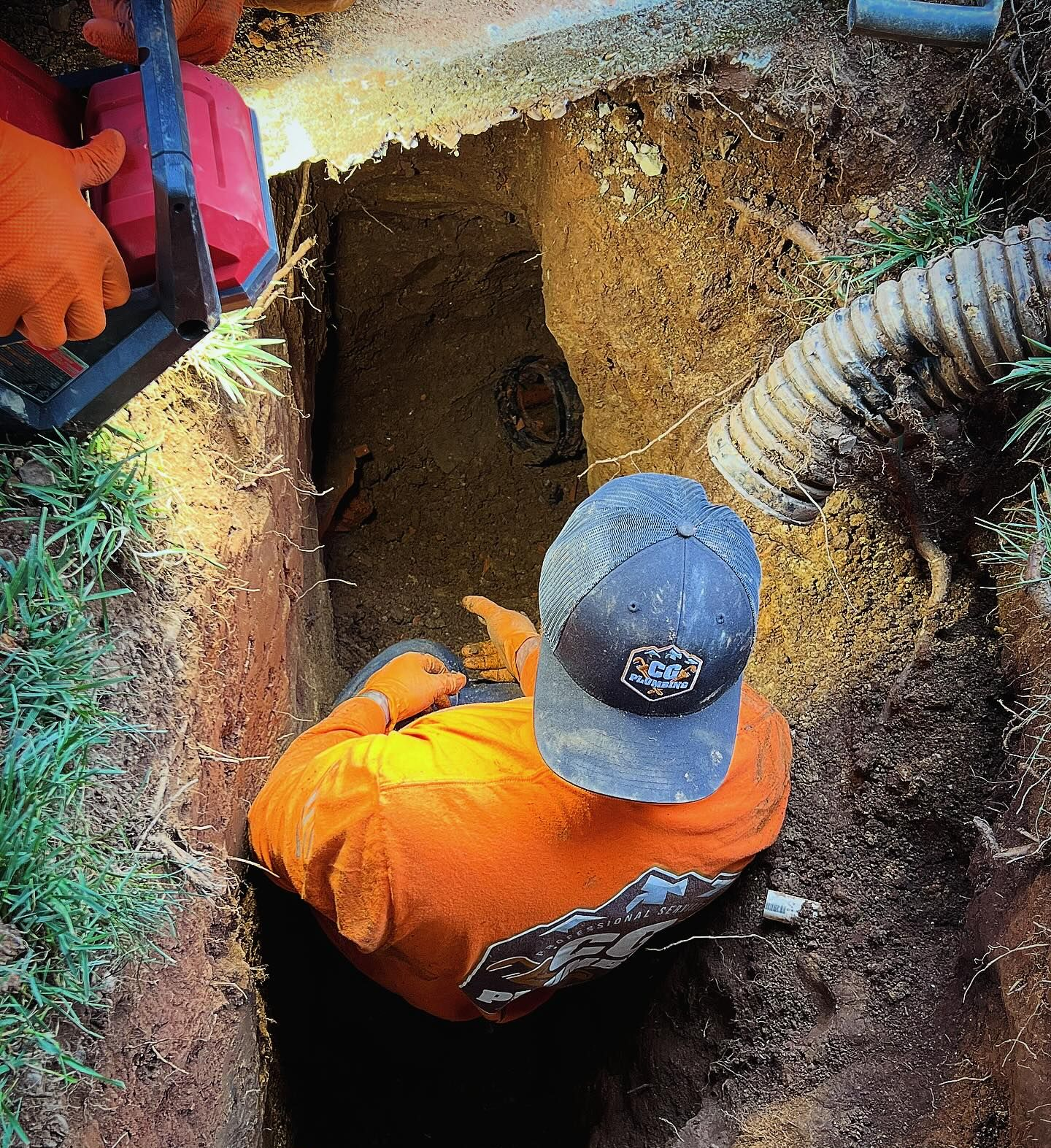 A person wearing an orange long-sleeved shirt and cap working in a narrow trench in the dirt, next to a drainage pipe.