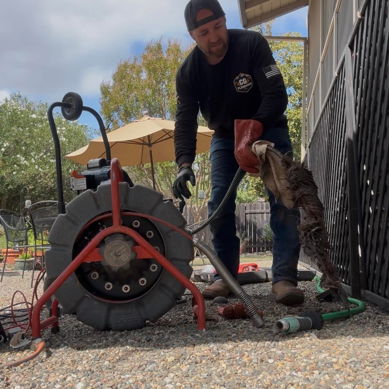 A person wearing gloves holds a mass of roots cleared from a pipe next to a red-and-black industrial drain auger outdoors.