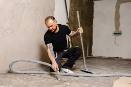 Worker using a floor tool to repair concrete in an unfinished room with exposed walls and piping