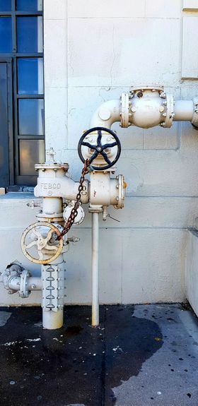 Industrial gas pipes and valves mounted on a white wall, with rust stains and wet ground below.