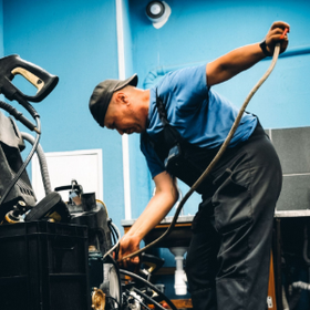 Mechanic in blue shirt bends over engine, using a hose in a workshop.
