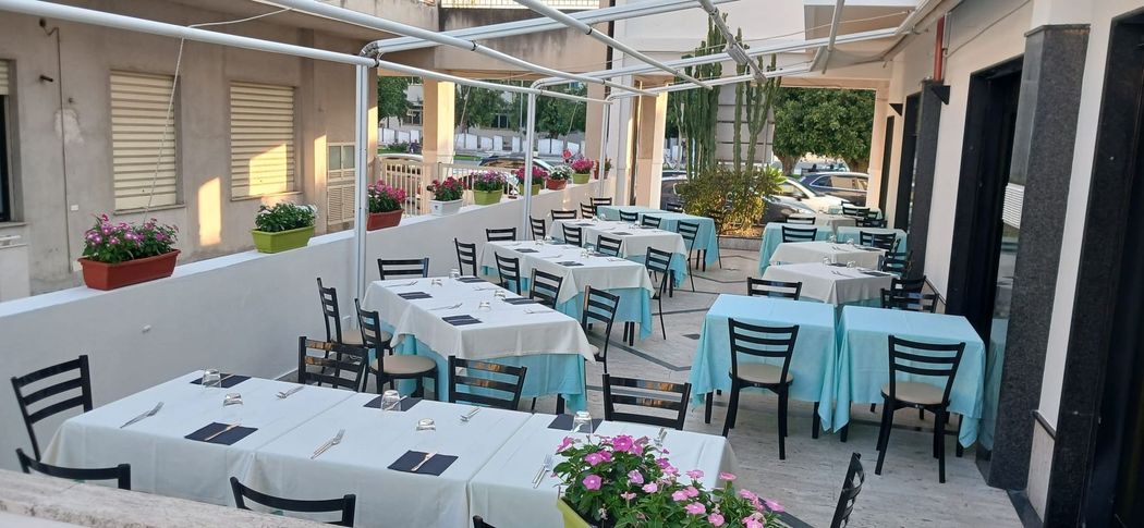 Area esterna con tavolini bianchi, sedie nere e tovaglie azzurre, sotto un patio coperto.