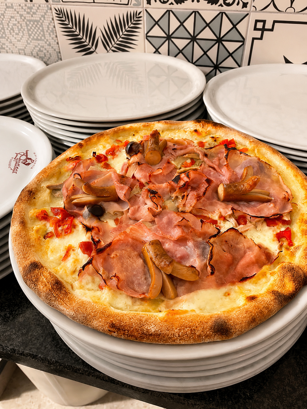 Pizza al prosciutto cotta al forno in una ciotola bianca su una pila di piatti, con piastrelle decorate sullo sfondo.