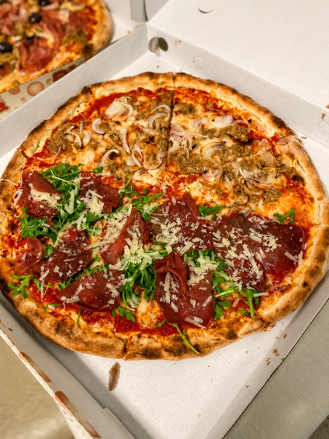 Pizza con peperoni e formaggio in una scatola aperta, guarnita con verdure ed erbe aromatiche, con una pizza parziale in alto a sinistra.