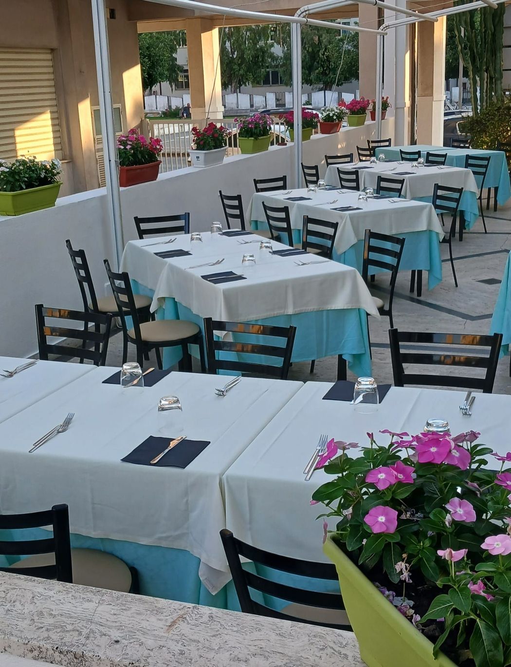 Dehor di un caffè all'aperto con tovaglie bianche, sedie turchesi e fiori rosa vicino a un pergolato.