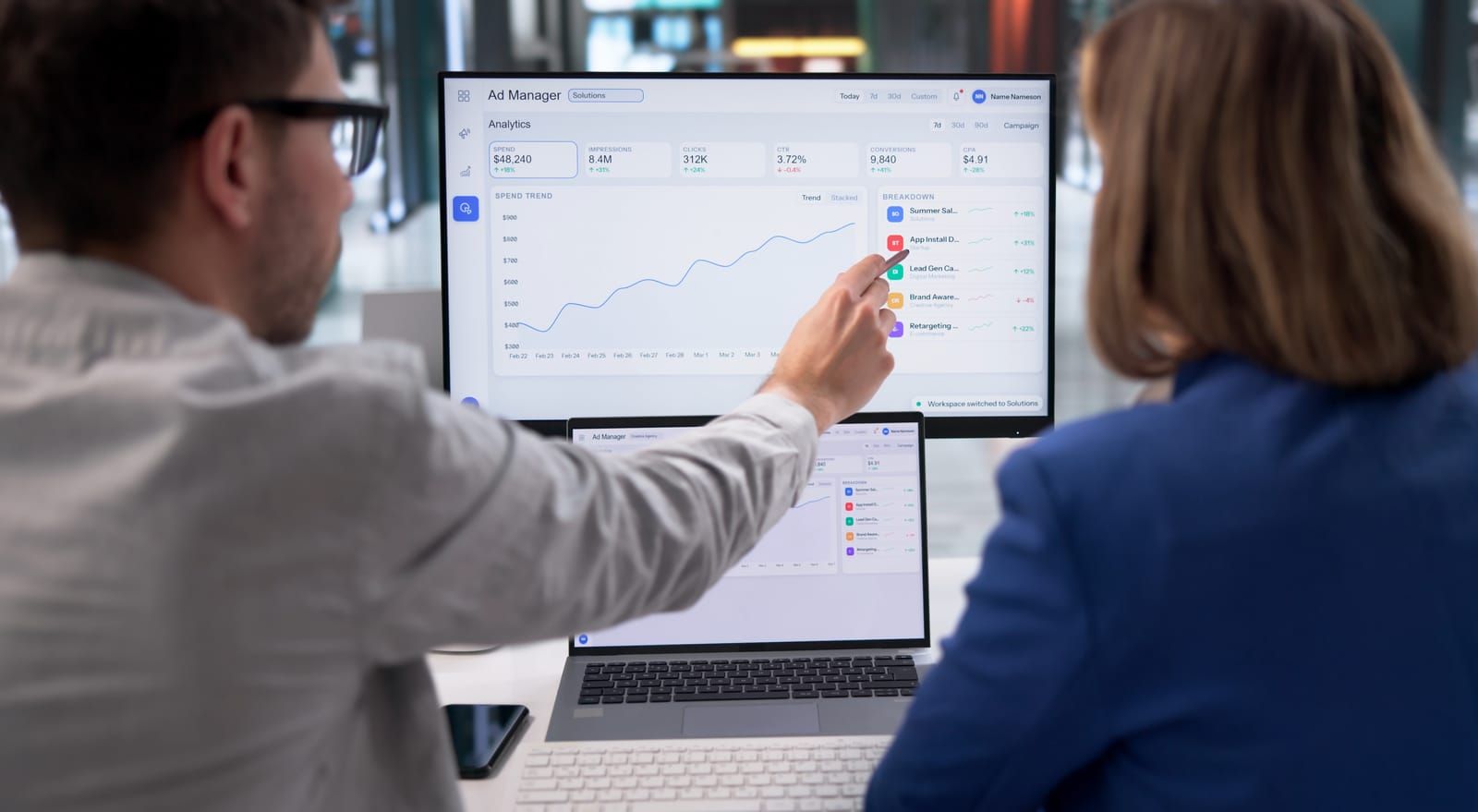 Two coworkers review a laptop dashboard with charts on a large monitor in an office.