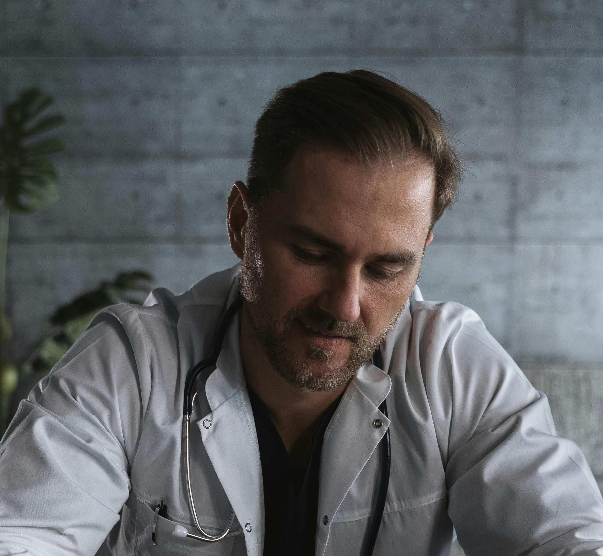 Doctor writing in a notebook at a table, wearing a light shirt in a dim room.
