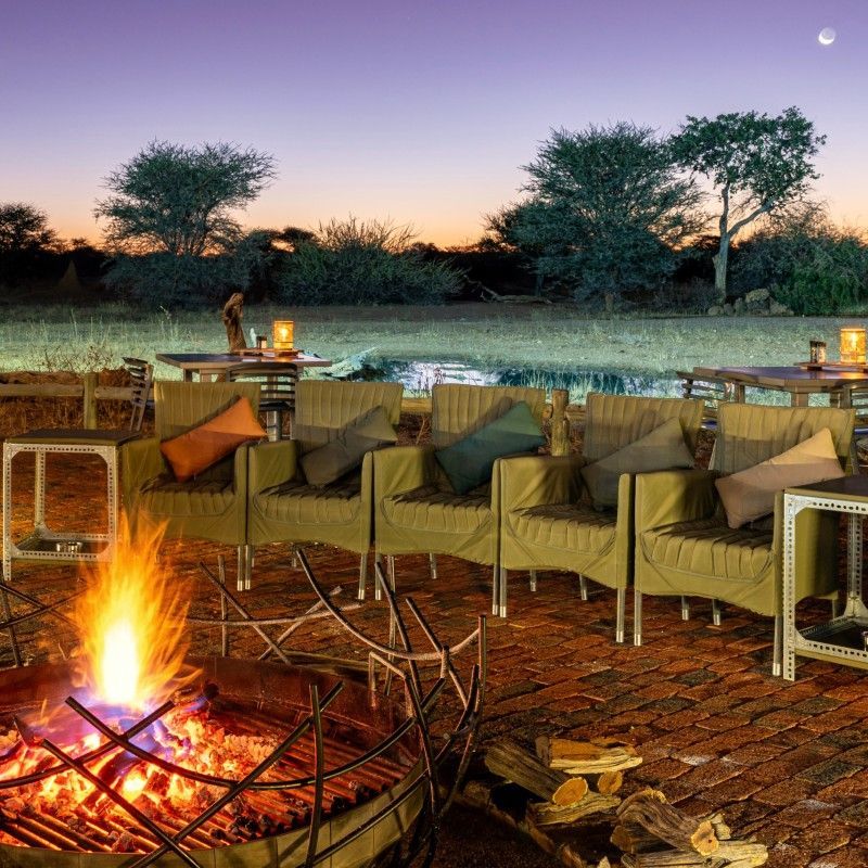 Camp chairs gathered around a glowing campfire at Okonjima Plains Camp in Namibia are captured in this atmospheric travel photograph, evoking the timeless spirit of an African safari evening. As the fire casts a warm, flickering light, the surrounding plains fade into the dusk, creating an intimate and inviting setting for storytelling, quiet reflection, and shared moments under the vast Namibian sky. This communal space reflects the camp’s relaxed elegance and deep connection to the wilderness, where guests come together after days spent exploring the reserve. Ideal for travelers seeking authenticity, connection, and unforgettable safari moments, this scene captures the warmth, tranquility, and magic of evenings at Okonjima Plains Camp. Inspiration Africa specializes in bespoke, tailor-made journeys to Namibia and across Africa, crafting immersive safari experiences and extraordinary moments in the heart of the African wilderness.