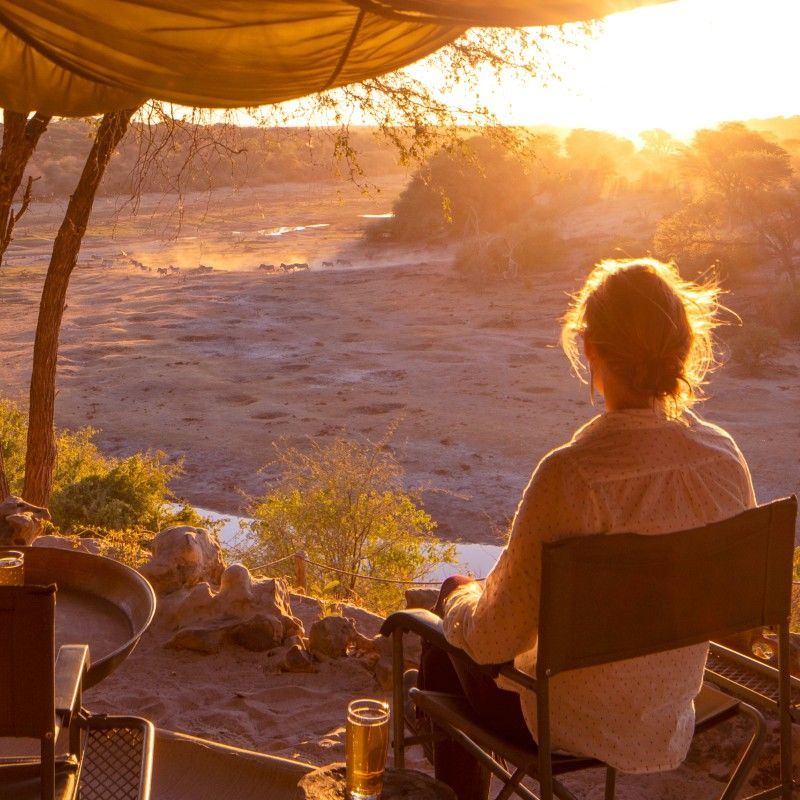 A guest overlooking the sunset from the balcony at Meno a Kwena in Botswana is captured in this atmospheric travel photograph, showcasing a moment of quiet reflection above the Boteti River. From the elevated viewpoint, warm golden light spreads across the riverbed and surrounding savannah, illuminating the landscape as the day draws to a close. The balcony setting offers an intimate connection to the natural environment, where wildlife is often seen moving along the river below. Natural materials and simple design frame the view without distraction, allowing the focus to remain on the dramatic colors of the sky and the vast openness of the setting. This scene reflects the timeless appeal of a Botswana safari, defined by space, serenity, and unforgettable sunsets. Inspiration Africa specializes in bespoke, tailor-made journeys to Botswana and across Africa, crafting unforgettable safari experiences in extraordinary wilderness destinations.
