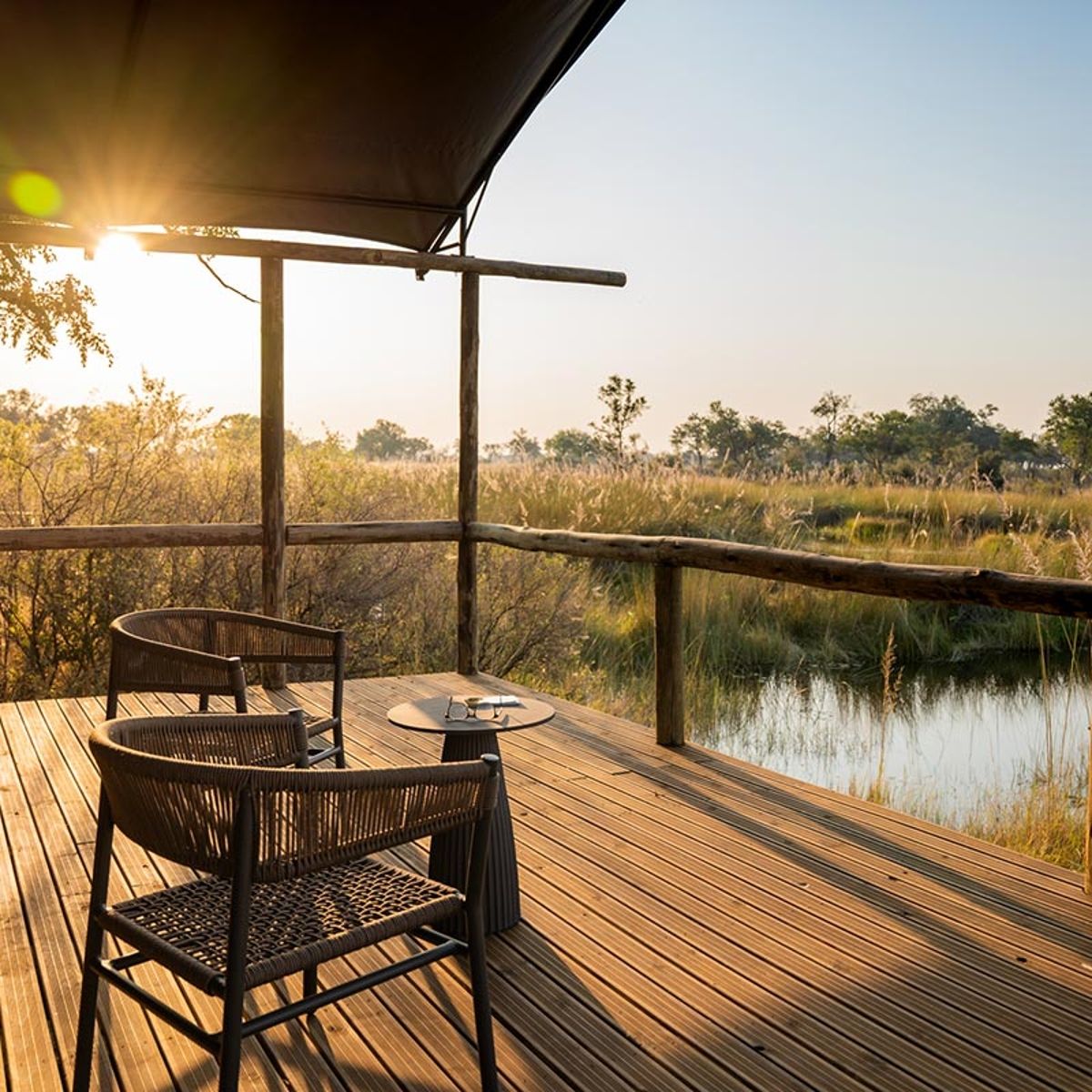 The veranda at 4 Rivers Camp by Kwando Safaris in Botswana is captured in this inviting travel photograph, showcasing a spacious, open-air deck overlooking the lush floodplains and waterways of the Kwando region. Comfortable seating and natural wooden furnishings create a relaxed space for guests to enjoy panoramic views of the surrounding wilderness, where elephants, antelope, and birds are often seen grazing or moving through the landscape. Soft lighting and the warm tones of the setting sun enhance the tranquil and immersive safari atmosphere. The scene conveys comfort, connection to nature, and the unique serenity of Botswana’s Okavango-adjacent wilderness, highlighting 4 Rivers Camp as a premier destination for authentic safari experiences. Inspiration Africa specializes in bespoke, tailor-made journeys to Botswana and across Africa, crafting unforgettable safari experiences in extraordinary destinations.