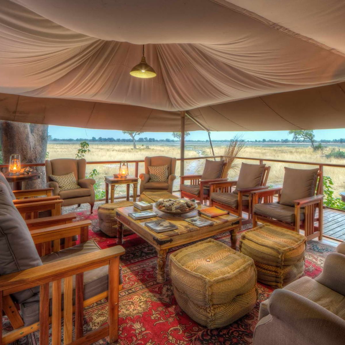 The outdoor dining area at Kadizora Camp by African Anthology in Botswana is captured in this atmospheric travel photograph, showcasing an elevated deck set amid the lush floodplains of the Okavango Delta. Tables are arranged to maximize views of the surrounding waterways, where wildlife such as elephants, hippos, and birds often pass by, creating a dynamic and immersive dining experience. Natural wooden furnishings and shaded areas blend seamlessly with the environment, while warm lighting adds a welcoming and intimate atmosphere at dusk. The scene conveys elegance, comfort, and a deep connection to Botswana’s wilderness, highlighting Kadizora Camp as a premier safari destination where meals are shared in harmony with nature. Inspiration Africa specializes in bespoke, tailor-made journeys to Botswana and across Africa, crafting unforgettable safari experiences in extraordinary destinations.