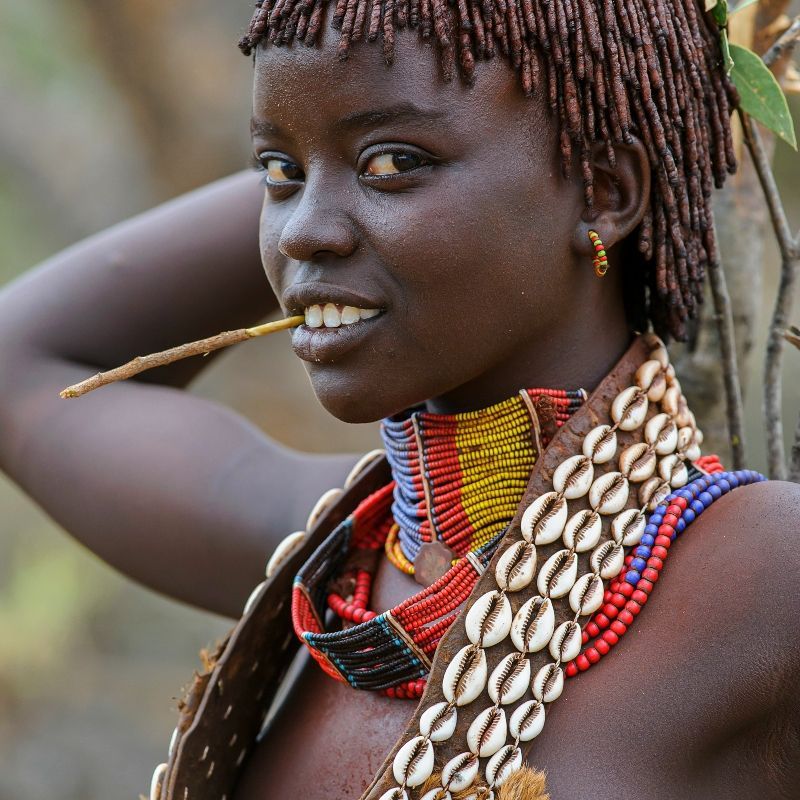 A Hamer woman in traditional attire in Ethiopia is captured in this striking travel photograph, highlighting the rich cultural heritage and distinctive identity of one of the country’s most well-known ethnic communities. Adorned with layered beads, metal jewelry, and intricately styled ochre-colored hair, she stands against the backdrop of Ethiopia’s remote southern landscapes. The detailed textures of her clothing and adornments reflect generations of cultural tradition and craftsmanship, while soft natural light enhances the warmth and authenticity of the scene. The image conveys dignity, individuality, and a strong connection to cultural heritage. Inspiration Africa curates bespoke, tailor-made journeys to Ethiopia and across Africa, combining immersive cultural encounters with authentic travel experiences in extraordinary destinations.