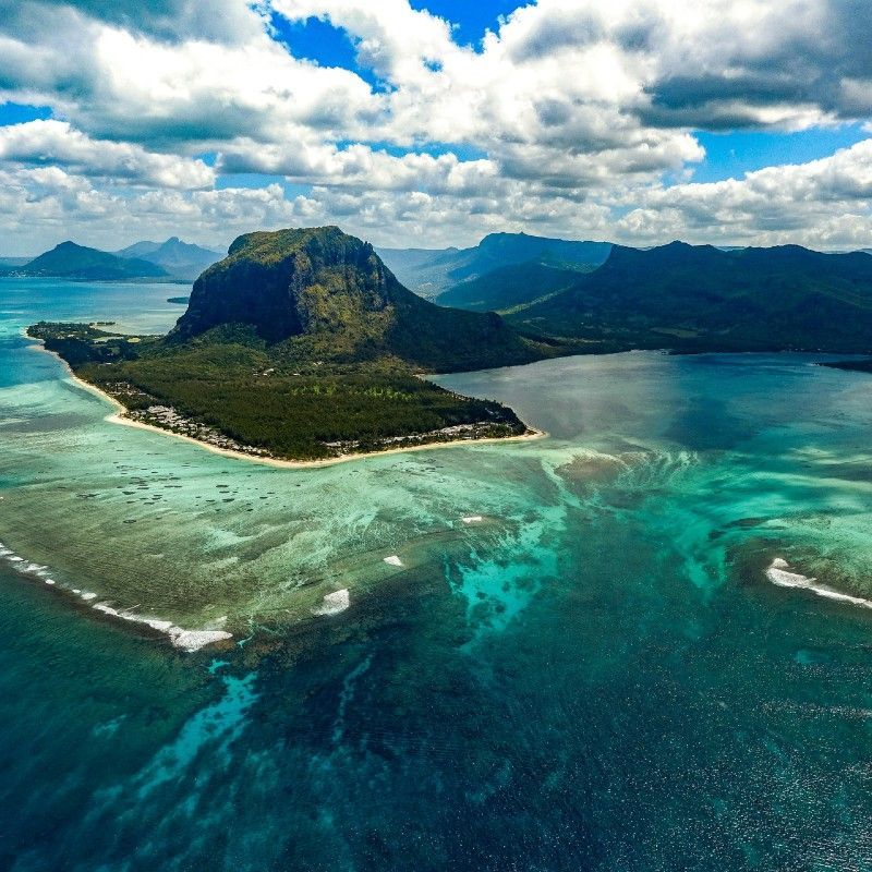 Witness the awe-inspiring Le Morne Underwater Waterfall in Mauritius from the air. This aerial shot captures the mesmerizing optical effect, where varying shades of ocean water—from deep sapphire blue to bright turquoise—create the illusion of a flowing cascade. The majestic Le Morne mountain, draped in lush green vegetation, dramatically anchors the scene. Inspiration Africa designs your ultimate, tailor-made trip to Mauritius and other exceptional African destinations. Explore this geological wonder and book your dream island getaway with us!